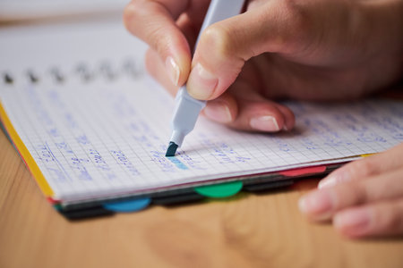 Woman using highlighter marker on squared notebook paper, closeup of right hand holding pen and making notes, visible fingernails and part of palm, wooden desk backgroundの写真素材