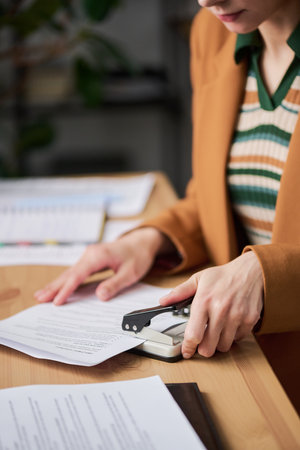 Caucasian young adult woman using paper puncher at desk, focusing on paperwork with hands visible, performing office task in professional workspace, not looking at cameraの写真素材