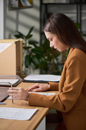 Young adult Caucasian woman working at desk reviewing documents and making notes, sitting in modern office environment with paperwork and folders organized on workspaceの写真素材