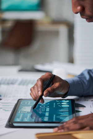 Black man using stylus interacting with digital tablet displaying data charts and analytics, close up of hand and device on desk, adult engaged in technology related taskの写真素材