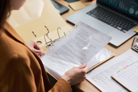 Young adult woman holding document in transparent sheet protector, reviewing paperwork at desk with open binder, laptop and notebook, focusing on business or administrative taskの写真素材