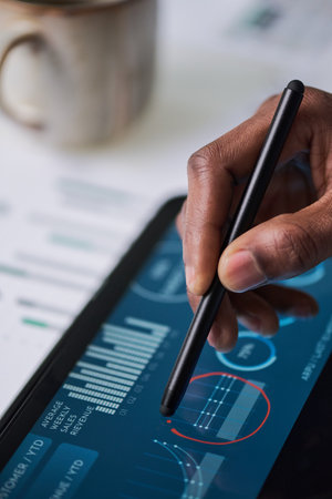 Black man using digital pen interacting with tablet displaying financial charts and analytics, hand holding stylus making notes on touchscreen, closeup of technology in business settingの写真素材