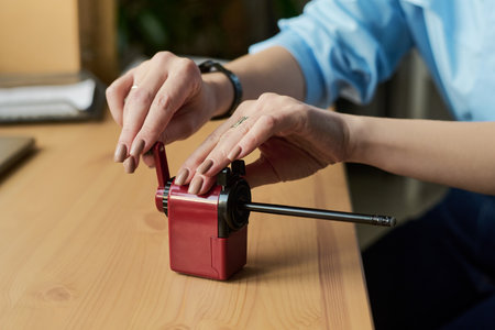 Young adult woman sharpening pencil using manual pencil sharpener on wooden desk, hands visible adjusting crank handle, wearing wristwatch, natural nails, close upの写真素材
