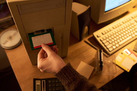 Caucasian young adult hand inserting labeled floppy disk into vintage desktop computer, surrounded...