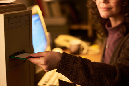 Caucasian woman inserting floppy disk into vintage desktop computer, sitting at desk with monitor in...