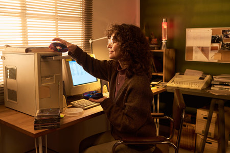 Caucasian woman sitting at desk using floppy disks with vintage computer equipment, holding disk in...