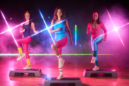 Three young adult women performing aerobic step exercises on platforms in fitness studio, smiling...