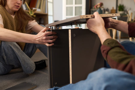 Caucasian young woman and Caucasian young man assembling furniture together on floor, both using...