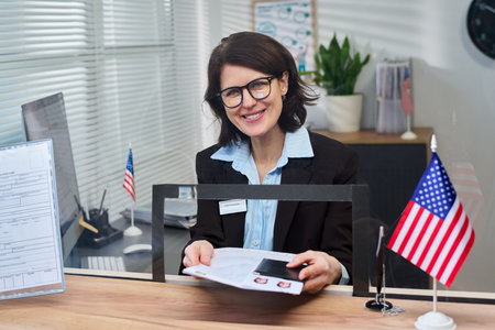 Portrait of middle aged woman sitting at visa center desk smiling while holding documents and...