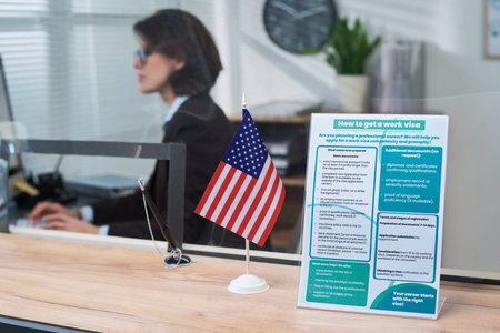 Caucasian woman working at visa center desk using computer, American flag and informational work visa brochure standing on counter in foreground, office environment visible in backgroundの写真素材