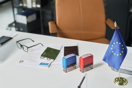 Visa center desk showing European Union flag, two passport stamps, eyeglasses, green passport, application forms with photo, processing travel documents for identification purposesの写真素材