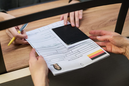Caucasian woman handing over visa application form and passport to office worker at visa center, both sets of hands visible, office worker holding pen and reviewing documentsの写真素材