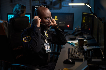 Black man wearing headset working at computer in security control room, monitoring multiple screens...