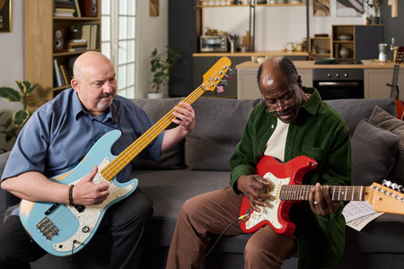 Middle aged Caucasian man and senior Black man sitting on sofa playing electric guitars together,...