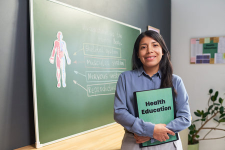 Portrait of young adult Hispanic woman standing in classroom holding Health Education folder,...