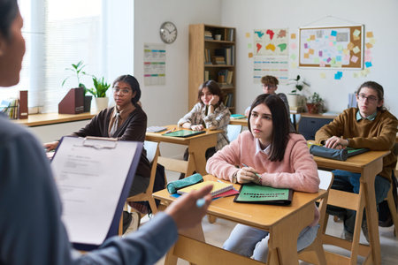 Group of multiethnic teenagers sitting at desks listening to teacher holding clipboard in classroom,...