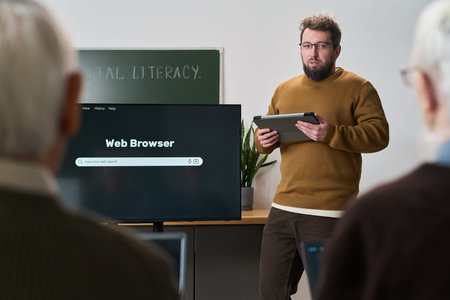 Caucasian young adult man holding digital tablet teaching computer literacy course to group of...