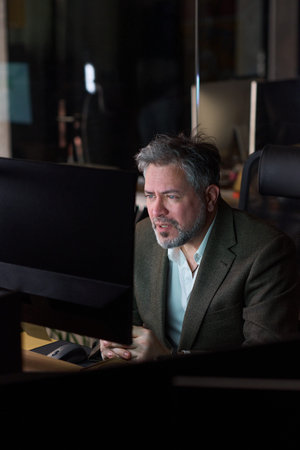 Middle aged Caucasian man sitting at desk working on computer monitor in modern office, facial hair visible, focused expression, hands clasped together, looking at screenの写真素材