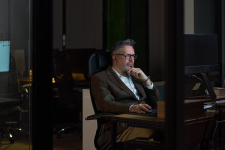 Middle aged Caucasian man sitting at desk working on computer in modern office at night, wearing...