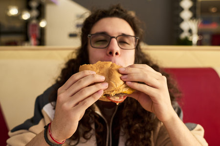 Gen Z man with long curly hair and glasses enjoying eating hamburger at restaurant, holding sandwich...