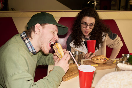 Young man eating sandwich while sitting at table with male friend drinking from red cup, both...