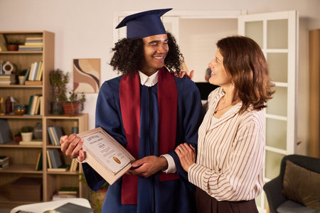 Young adult graduate holding diploma, smiling with middle aged mother at home, celebrating academic achievement and family support. Ideal for education success, parenting, future planning