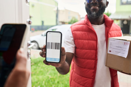 African American courier holding smartphone with barcode for scanning while delivering package. Image represents contactless delivery technology, ideal for e-commerce marketing