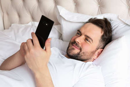 Handsome young man in bed typing on cell phone. Smiling caucasian man using mobile phone, browsing...