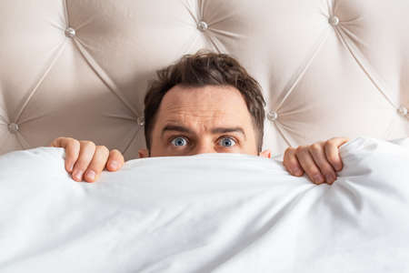 Caucasian young man hiding in bed under the blanket at home. He is stressed and tries to run away from problems.の写真素材