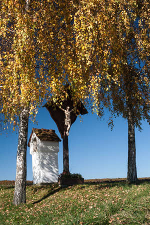 A christian wayside shrine and a cross under two birch trees in autumnの写真素材