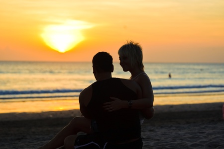 Two young lovers sitting on a beach and looking to each other on sunset backgroundの写真素材