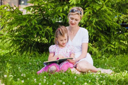 Little girl and her mother reading a book while they sitting and enjoy in beautiful natureの写真素材