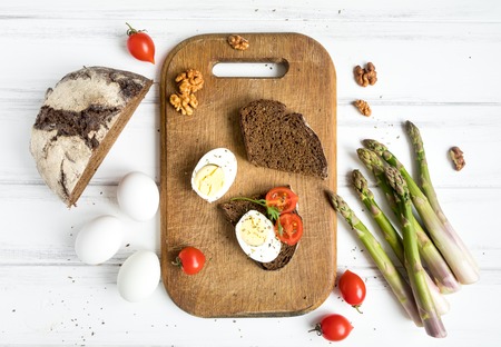 Fresh dark sliced bread, tomatoes, eggs on vintage wooden cutting board decorated with asparagus and nuts. Healthy rustic breakfast or snacks cooking. Flat lay, top viewの写真素材