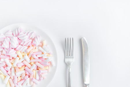 Colorful light marshmallows on a white plate with fork and knife on white background. Light soft...
