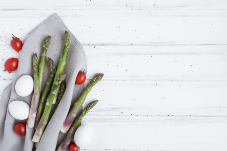 Ingredients for cooking healthy dietary breakfast: fresh asparagus, tomatoes, eggs on white vintage wooden table. Rustic style composition of vegetables and eggs on linen cloth. Flat lay, top viewの写真素材