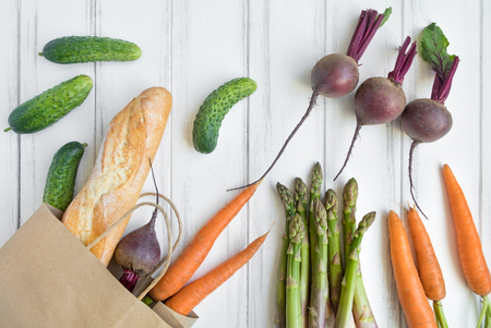 Paper shopping bag with fresh bread, carrots, cucumbers, asparagus and beet. Eco shopping concept, products for cooking. Flat lay on white vintage wooden table. Top view with place for textの写真素材