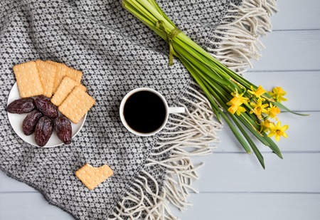 Cup of coffee, cracker biscuit cookies, dry date fruits and daffodil flowers. Cozy home composition on a plaid on wooden floor. Top view, flat layの写真素材