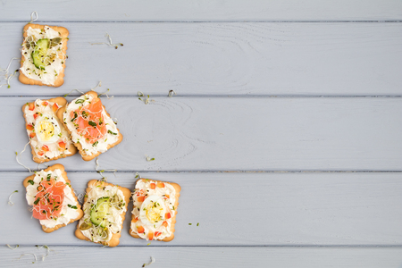 Frame of tasty crackers with cream cheese, salmon, eggs, cucumbers, seeds and micro greens. Appetizers on grey table. Healthy snacks, top view, flat lay, space for textの写真素材