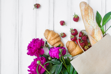 Paper shopping bag with fresh bread, croissants, strawberries, peony flowers. Eco shopping concept, bakery for breakfast. Flat lay on white vintage wooden table. Top view with place for textの写真素材