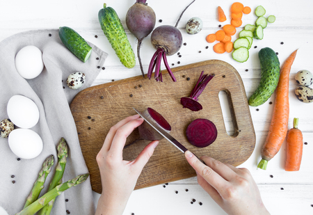 Woman hands slicing beetroot on wooden cutting board, surrounded by vegetables and eggs. Home cooking concept. Salad or any vegetarian dish. Flat lay, top viewの写真素材
