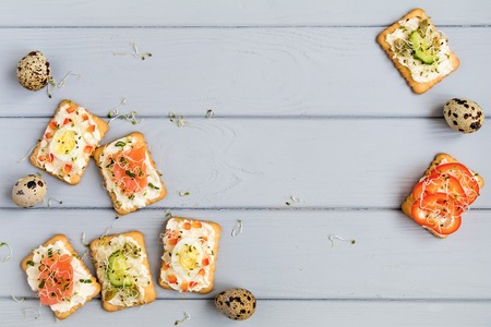 Frame of tasty crackers with cream cheese, sweet pepper, salmon, eggs, cucumbers, seeds and micro greens. Appetizers on grey table. Healthy snacks, top view, flat lay, space for textの写真素材