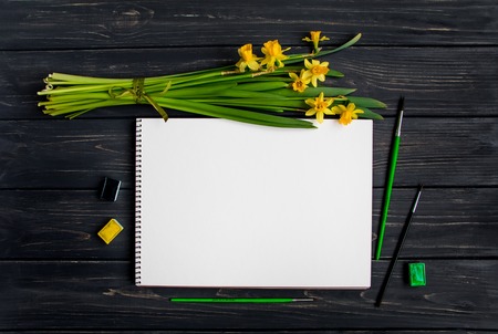 Natural composition with sketchbook, watercolor and brushes on black vintage wooden table, with bouquet of daffodils. Flat lay, top view, view from aboveの写真素材