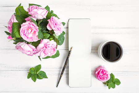 Composition with cup of coffee, sketchbook and rose flowers against white vintage wooden table. Cozy home morning composition. Sketching or writing and drinking coffee. Flat lay, top viewの写真素材