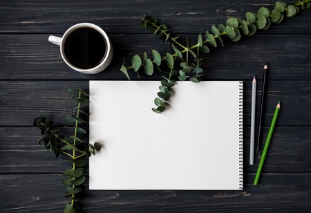 Natural composition with sketchbook, pencils and cup of coffee on black vintage wooden table, decorated with green eucalyptus branches. Flat lay, top view, view from aboveの写真素材