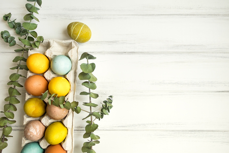 Easter eggs colored with colorful natural paints in an egg tray on white wooden table. Decorated with eucalyptus branches. Flat lay, top view with a space for textの写真素材