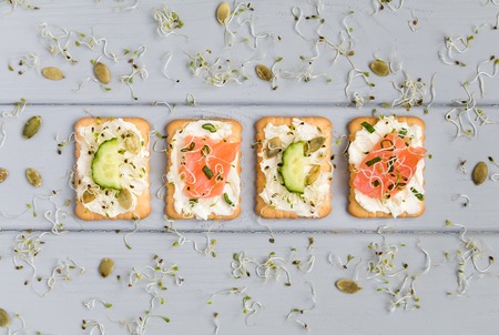 Tasty square crackers with cream cheese, cucumbers, salmon, pumpkin seeds and micro greens. Appetizers on grey table. Healthy vegetarian snacks, top view, flat layの写真素材