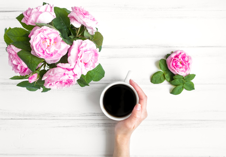 Woman hand holding cup of coffee surrounded with rose flowers against white vintage wooden table. Cozy home morning composition. Morning and drinking coffee. Flat lay, top viewの写真素材
