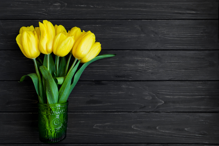 Beautiful bouquet of yellow tulips in green vintage glass vase on dark wooden table. Holiday gift or home decor concept. Top view, flat layの写真素材
