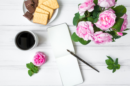 Composition with cup of coffee, sketchbook and rose flowers against white vintage wooden table. Cozy home morning composition with hot drink and sweets. Writing and drinking coffee. Flat lay, top viewの写真素材