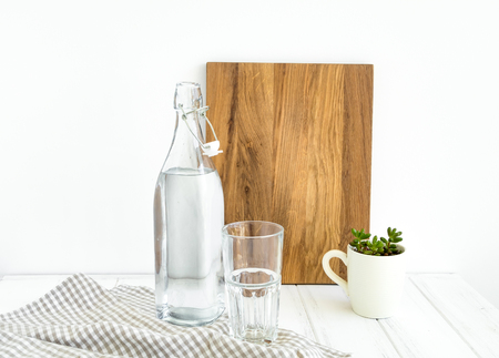 Vintage bottle of water, empty glass, plant in a cup, tablecloth and a wooden cutting board on white table, against white wall. Drinking clear water concept, minimalist home kitchen compositionの写真素材